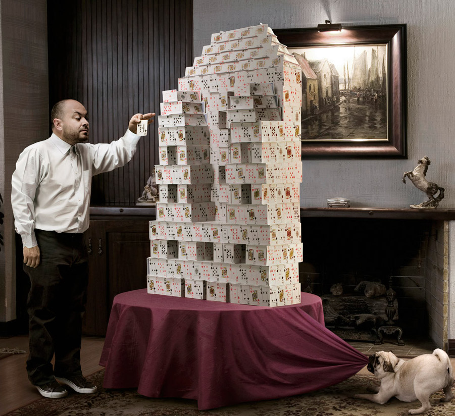 On a table, sitting on top of a tablecloth, is a large tower made out of playing cards. A man to the left is placing another card on top of the tower. To the right, a dog has the tablecloth in their teeth and is starting to pull